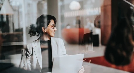 Smiling business woman in office on laptop seen through glass window 