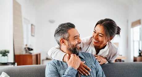  Smiling woman hugging her husband on the couch from behind in the living room. 