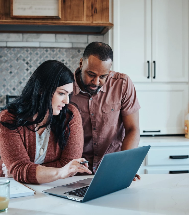Man and woman sit at kitchen counter with breakfast working with pen, paper and laptop.    