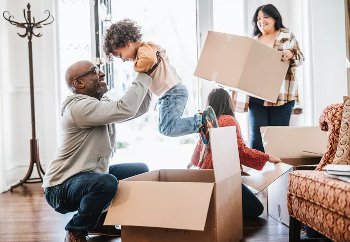 A bright indoor scene with several people unpacking moving boxes in a new home. In the foreground, a person kneels beside an open cardboard box and lifts a small child into the air. Another child sits near additional boxes, while a third person carries a large cardboard box through the doorway.