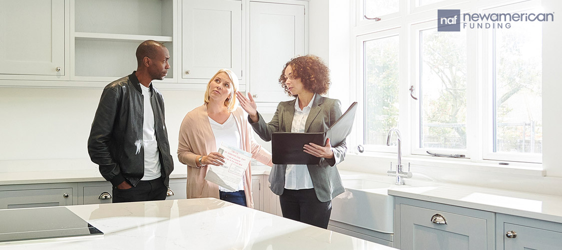 A couple stands in the kitchen of a home looking at a laptop with a professional