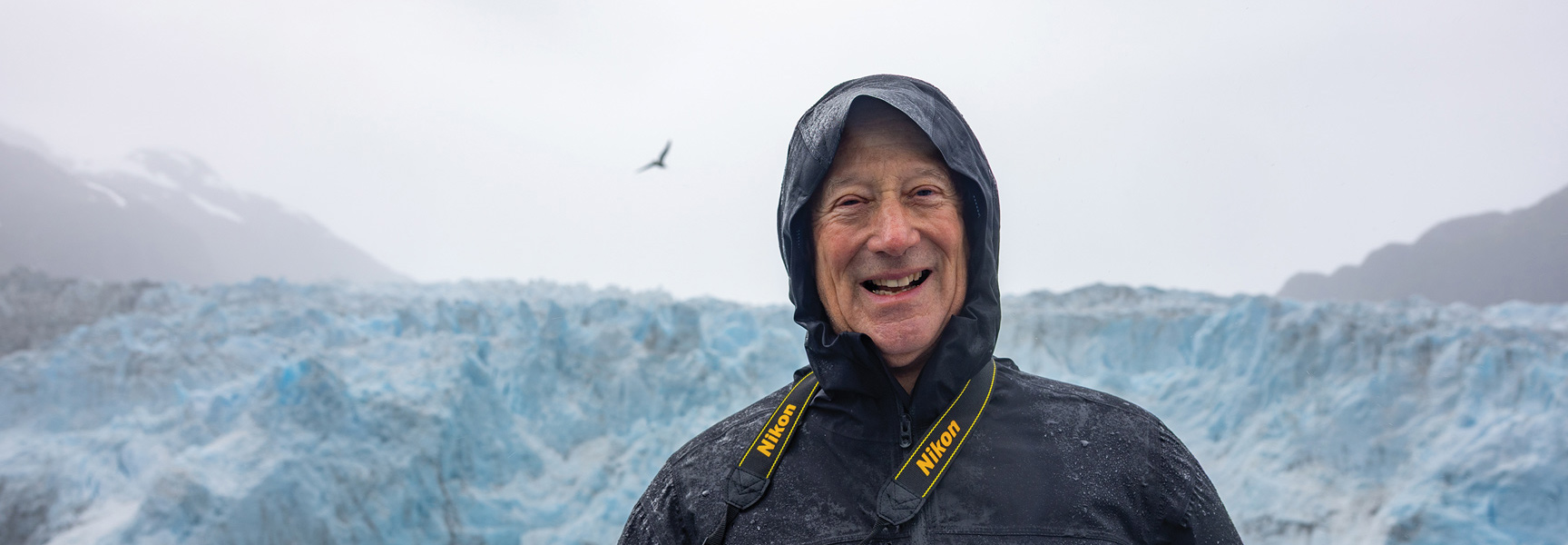 A smiling man in a black rain jacket stands in front of a massive blue glacier in Alaska.