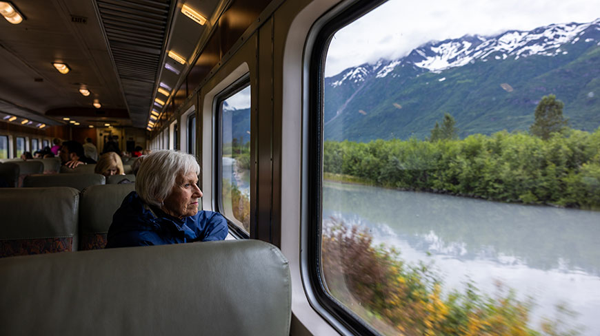 An older woman looks out a train window at a river and snow-capped mountains in Alaska.