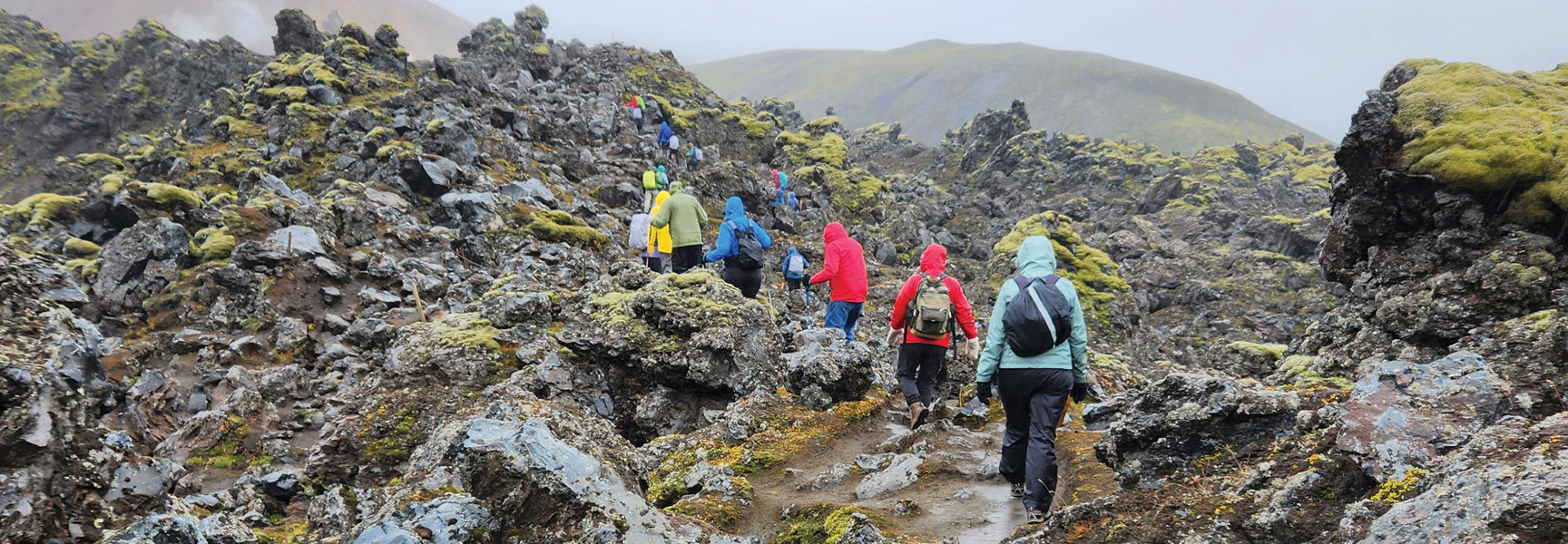 A group of hikers ascend a rocky, moss-covered trail in the dramatic volcanic landscape of South Iceland.