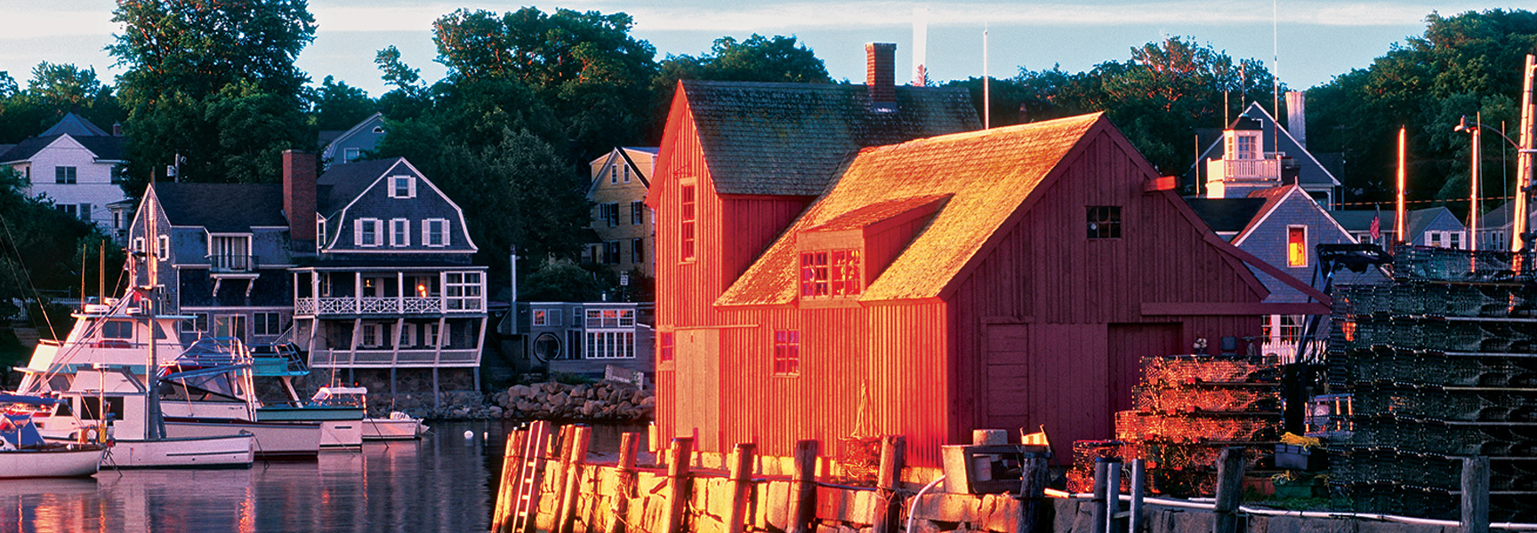 A red boathouse and classic New England homes line a harbor in Massachusetts during a golden sunset.