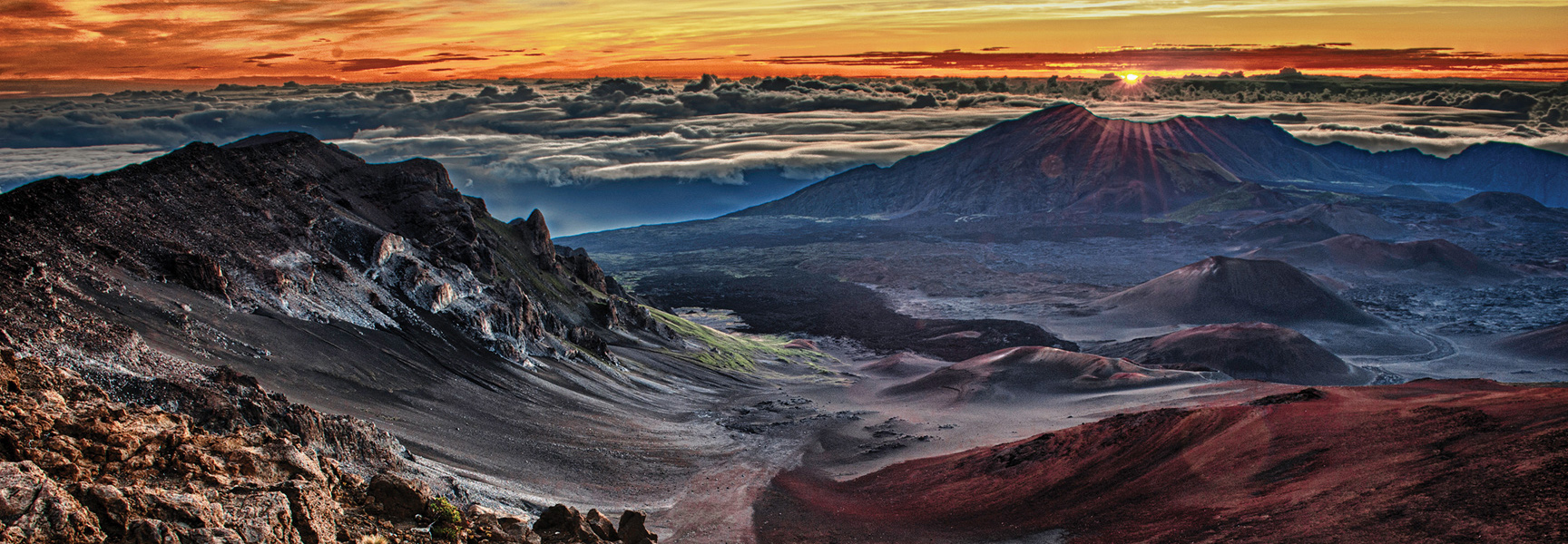 A dramatic sunrise over a vast volcanic crater in Hawaii, with rays of light cresting a distant peak above a sea of clouds.