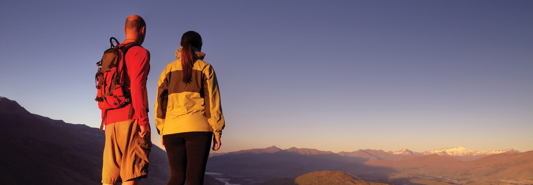 Two hikers at sunrise look out over a vast mountain range with snow-capped peaks in New Zealand.