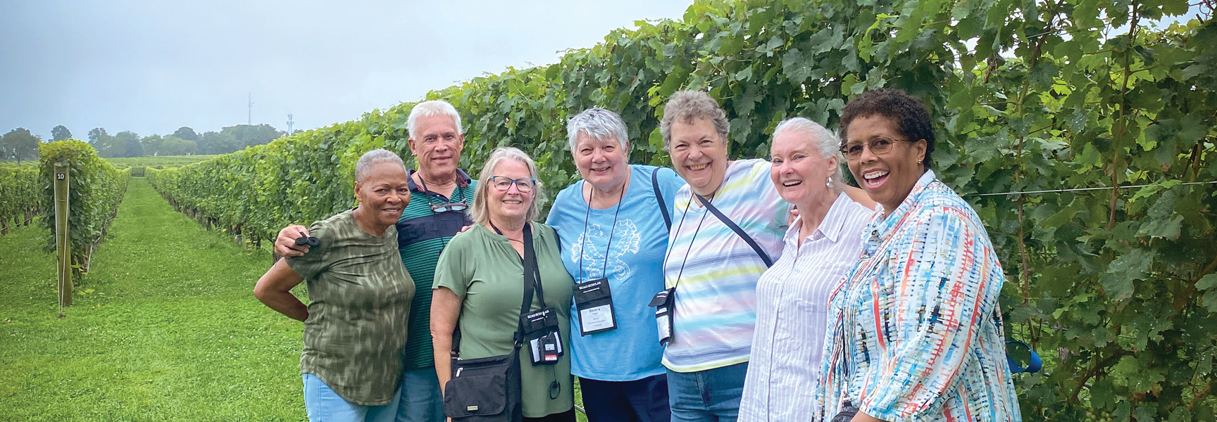 A group of seven older adults smile together for a photo in a lush, green New England vineyard.