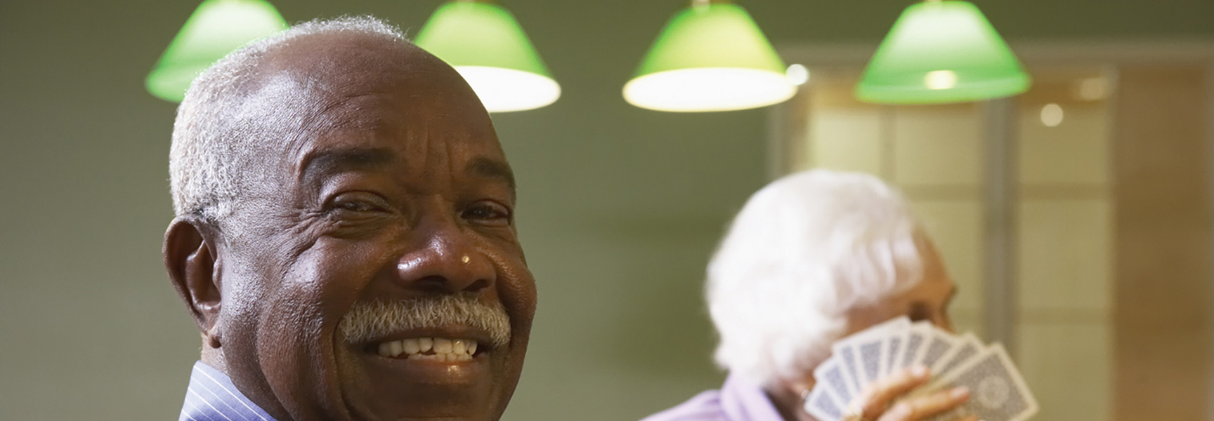 An older Black man smiles warmly during a bridge game in Georgia, with another player holding cards visible in the background.