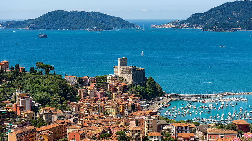 The colorful coastal village of Lerici, Italy, with its historic castle overlooking a harbor filled with boats on the bright blue Mediterranean Sea.
