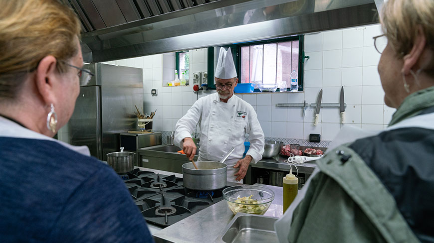 An Italian chef in a white uniform gives a cooking demonstration to two women in a professional kitchen in Italy.