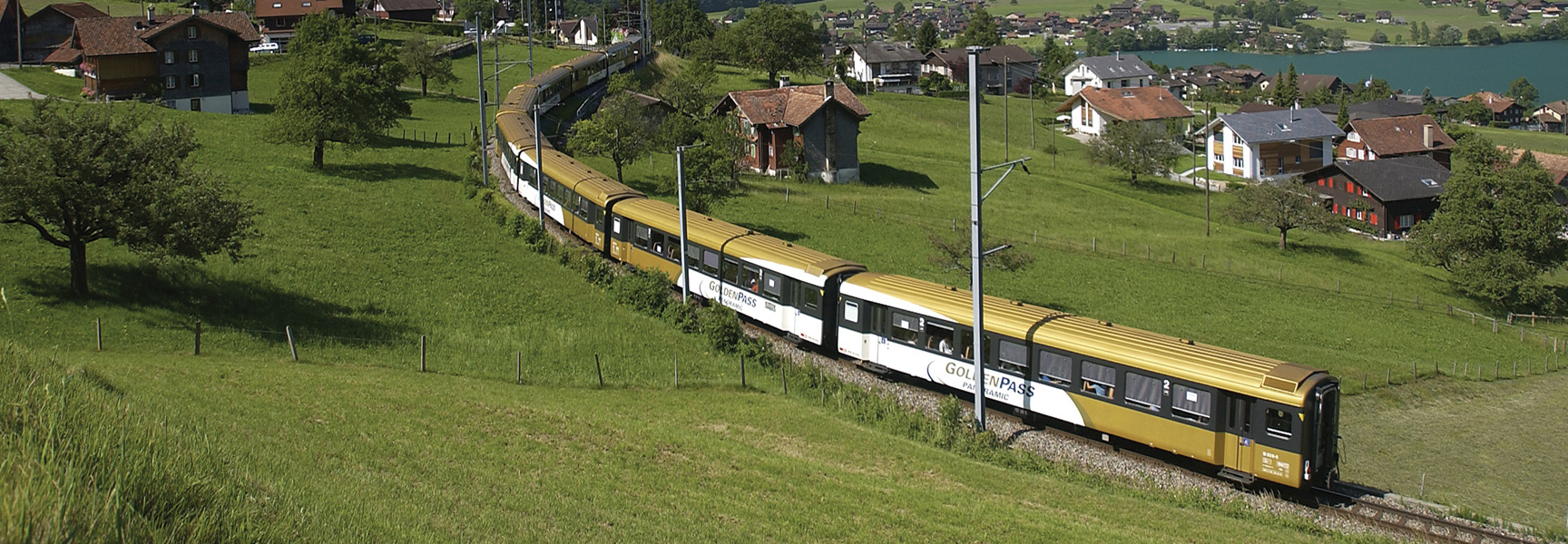 A golden train travels through the rolling green hills of a Swiss village next to a lake.