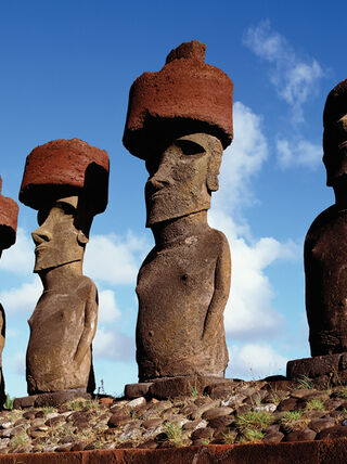 Five ancient moai statues stand against a blue sky on Easter Island (Rapa Nui), with four wearing large red stone topknots.