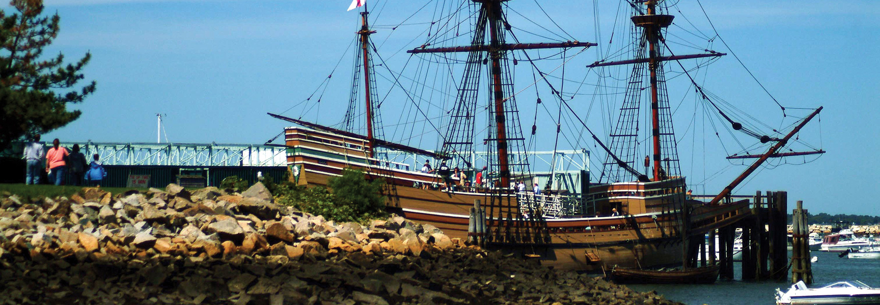 The Mayflower II replica ship is docked in Plymouth Harbor, Massachusetts, with a rocky shoreline in the foreground and visitors on deck.