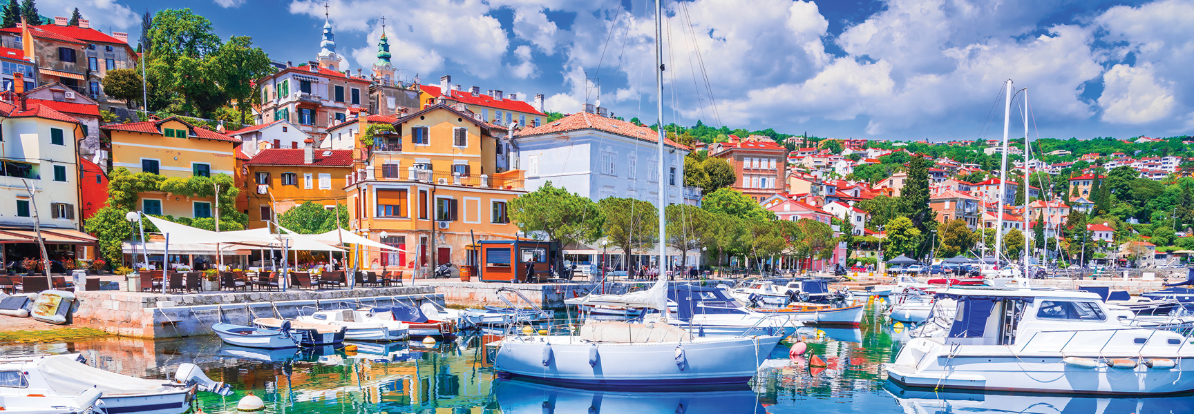 Sailboats and yachts are docked in a harbor in front of a colorful seaside village in Croatia.