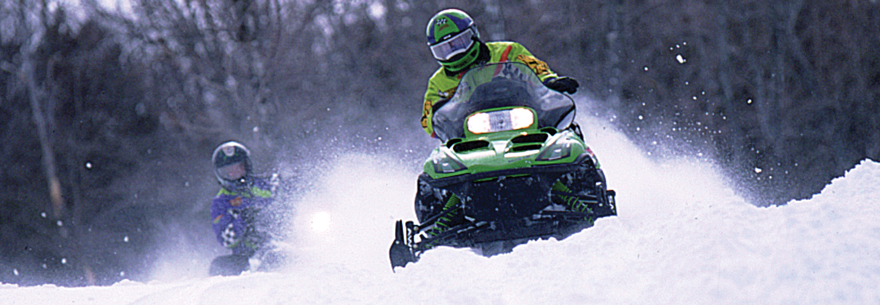Two people on snowmobiles kick up fresh powder while riding through a snowy forest during winter in Montana.