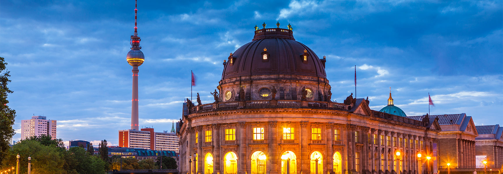 The Bode Museum in Berlin, Germany, is illuminated at dusk with the iconic TV Tower visible in the background against a blue sky.