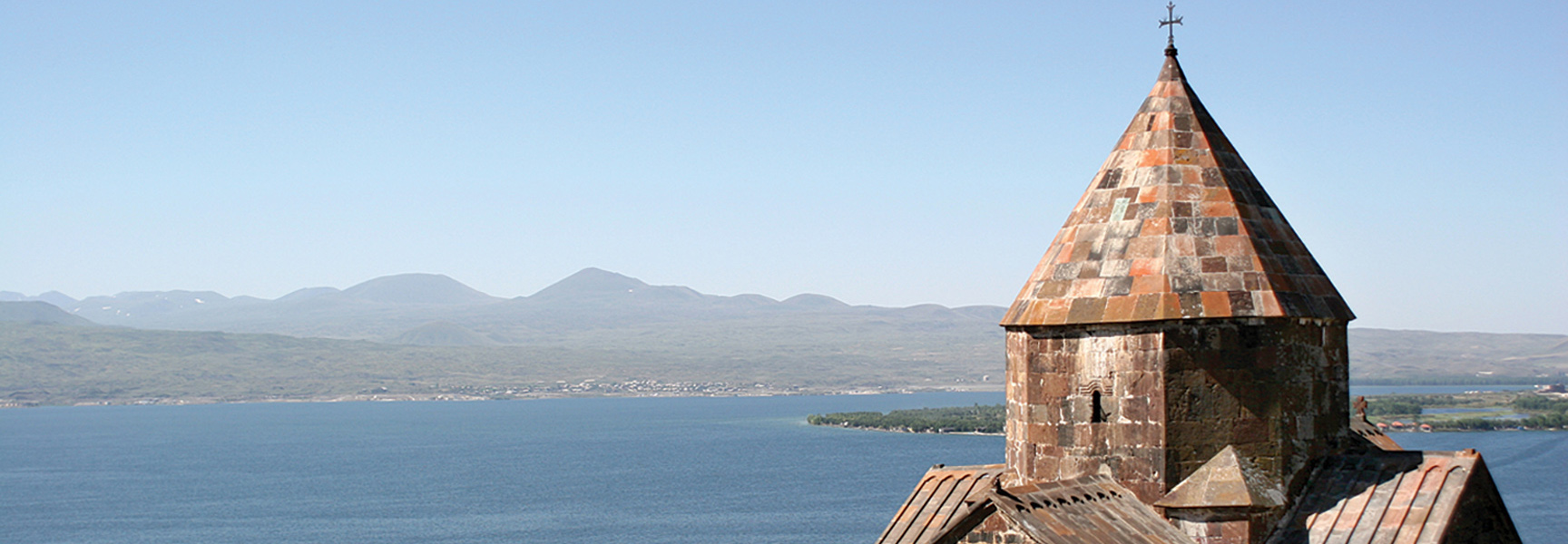 A stone church tower with a conical roof overlooks a large lake and a mountainous shoreline in the South Caucasus.