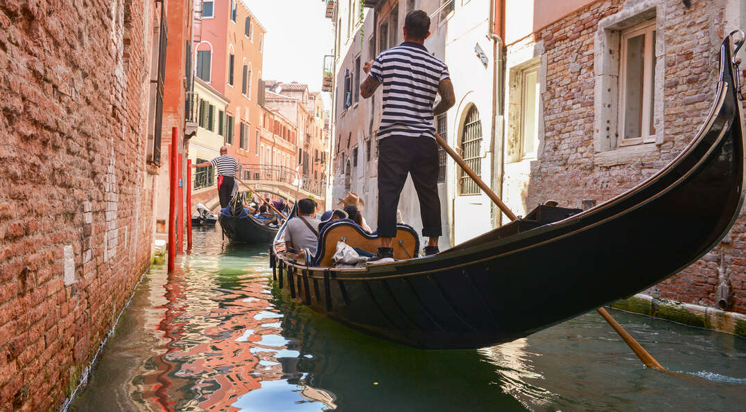 A gondolier in a striped shirt guides a gondola through a narrow canal lined with brick buildings in Venice, Italy.