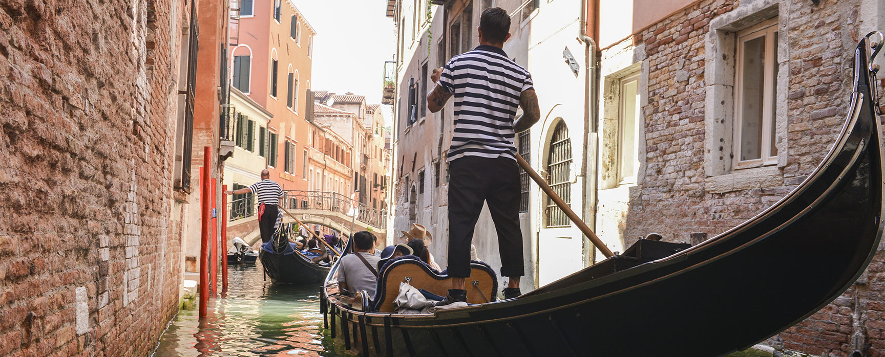 A gondolier steers a gondola with passengers down a narrow canal between tall brick buildings in Venice, Italy.