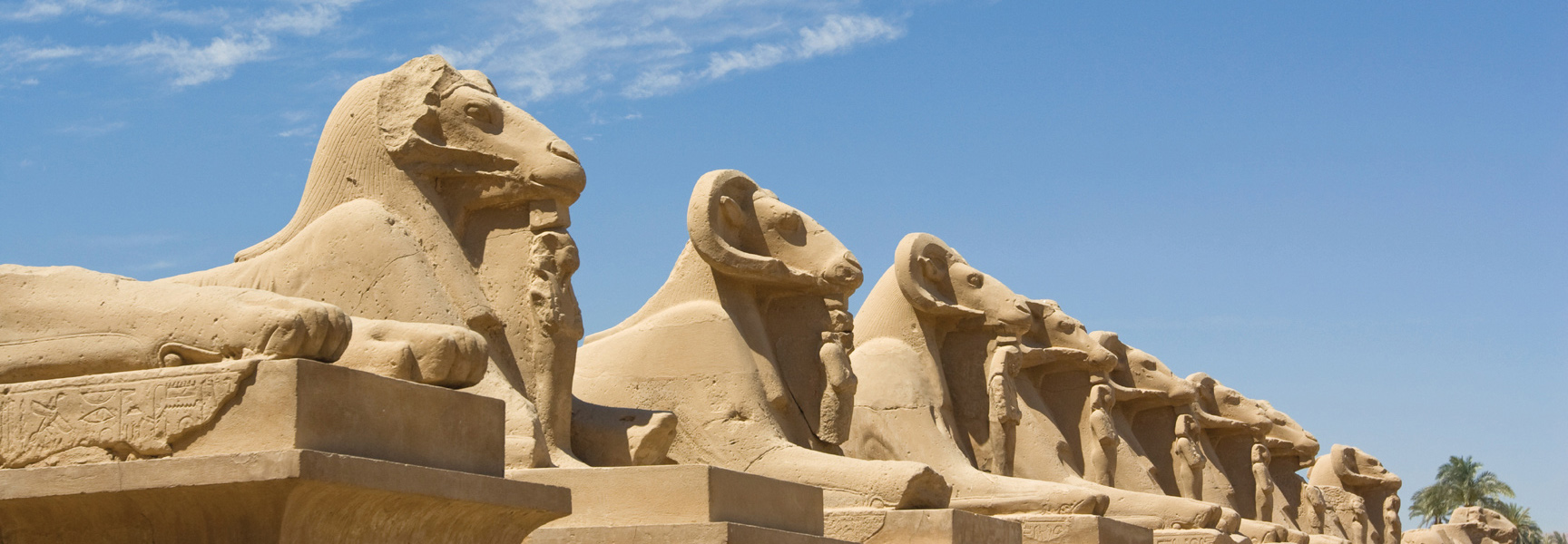 A row of ancient ram-headed sphinx statues line a path under a clear blue sky in Egypt.
