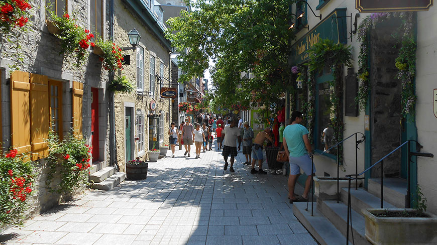 Pedestrians stroll along a sunlit Québec City street lined with historic stone buildings and flowers, capturing the area’s Old World charm and architecture.