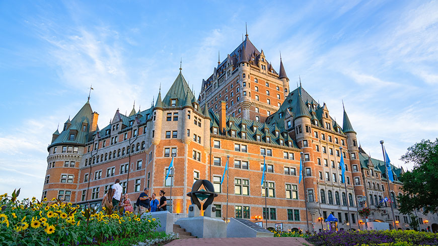 Tourists walk past the historic architecture of Old City Québec, featuring the iconic Château Frontenac under a blue sky with sunflowers in the foreground.