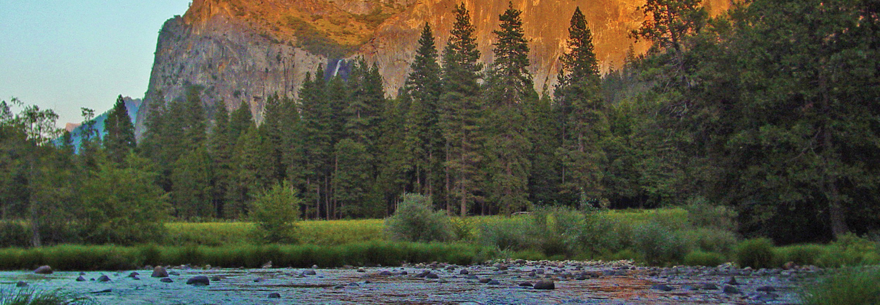 A rocky river flows in front of a pine forest and large granite cliff face at sunset in Yosemite National Park, California.