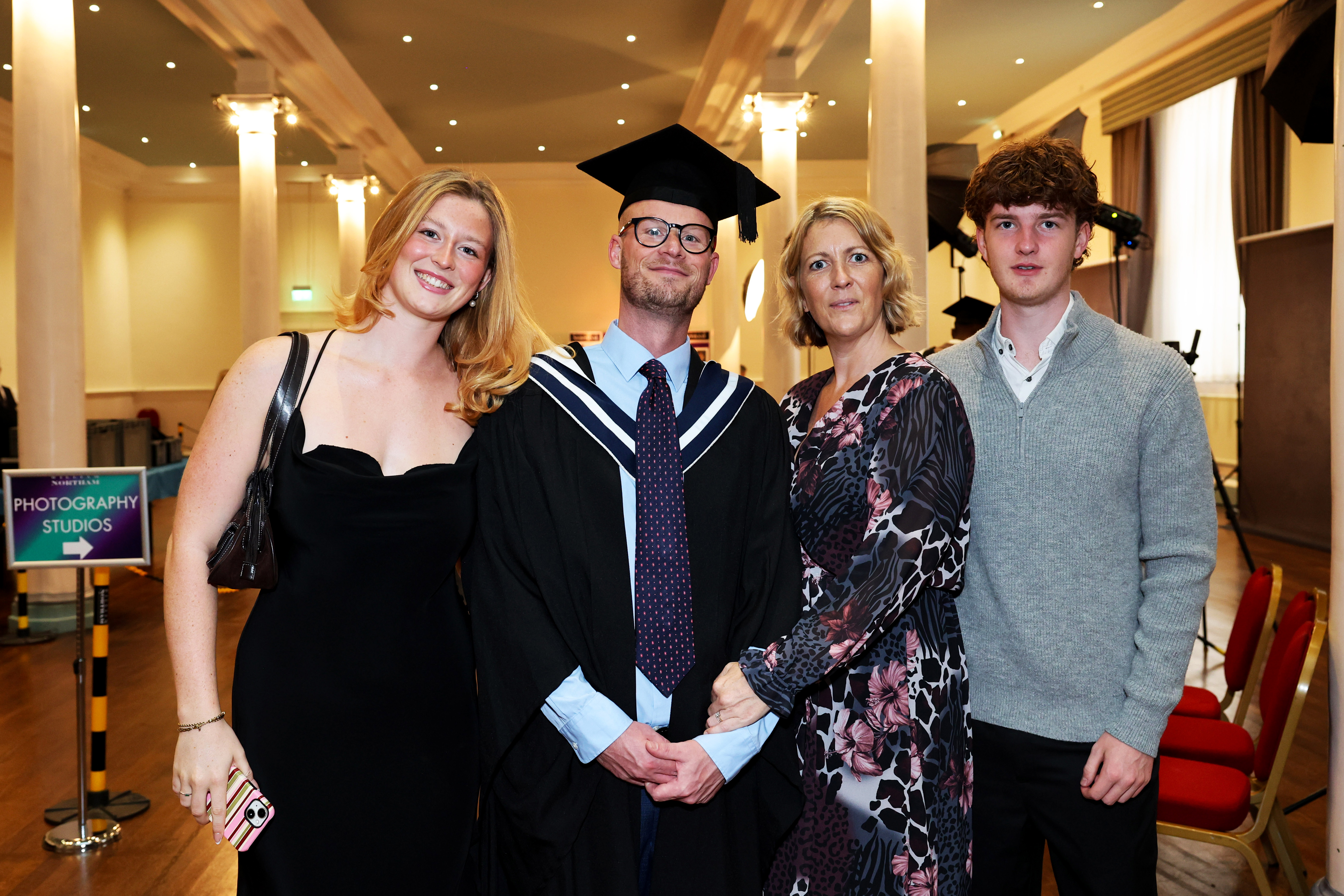 Male graduate in cap and gown posing, surrounded by 3 family members