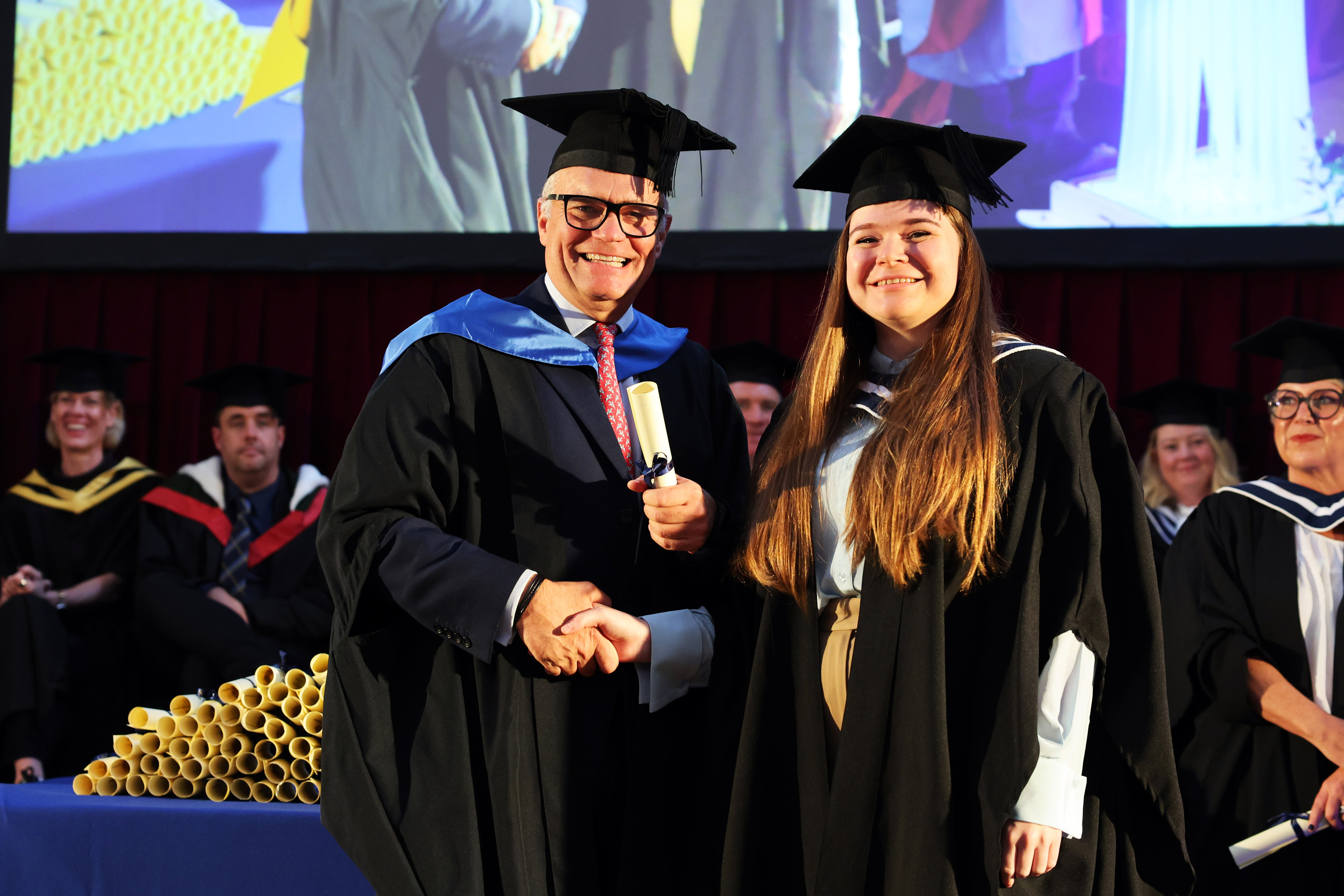 A female graduate on stage shaking hands and receiving a scroll from Ben Farrell, CIPS CEO