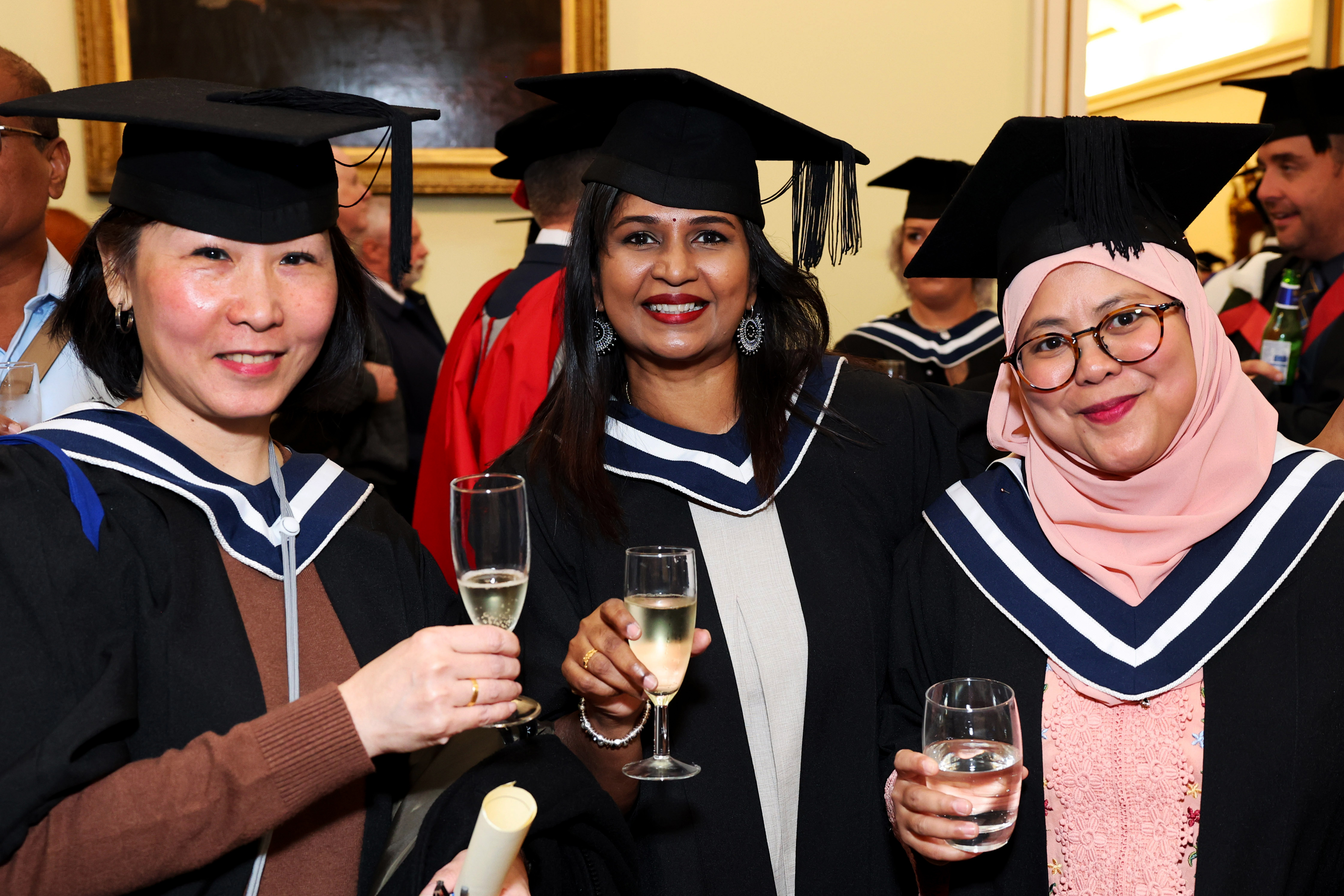 Three women of mixed ethnicities in graduation gap and gown pose side by side holding drinks at the graduation reception
