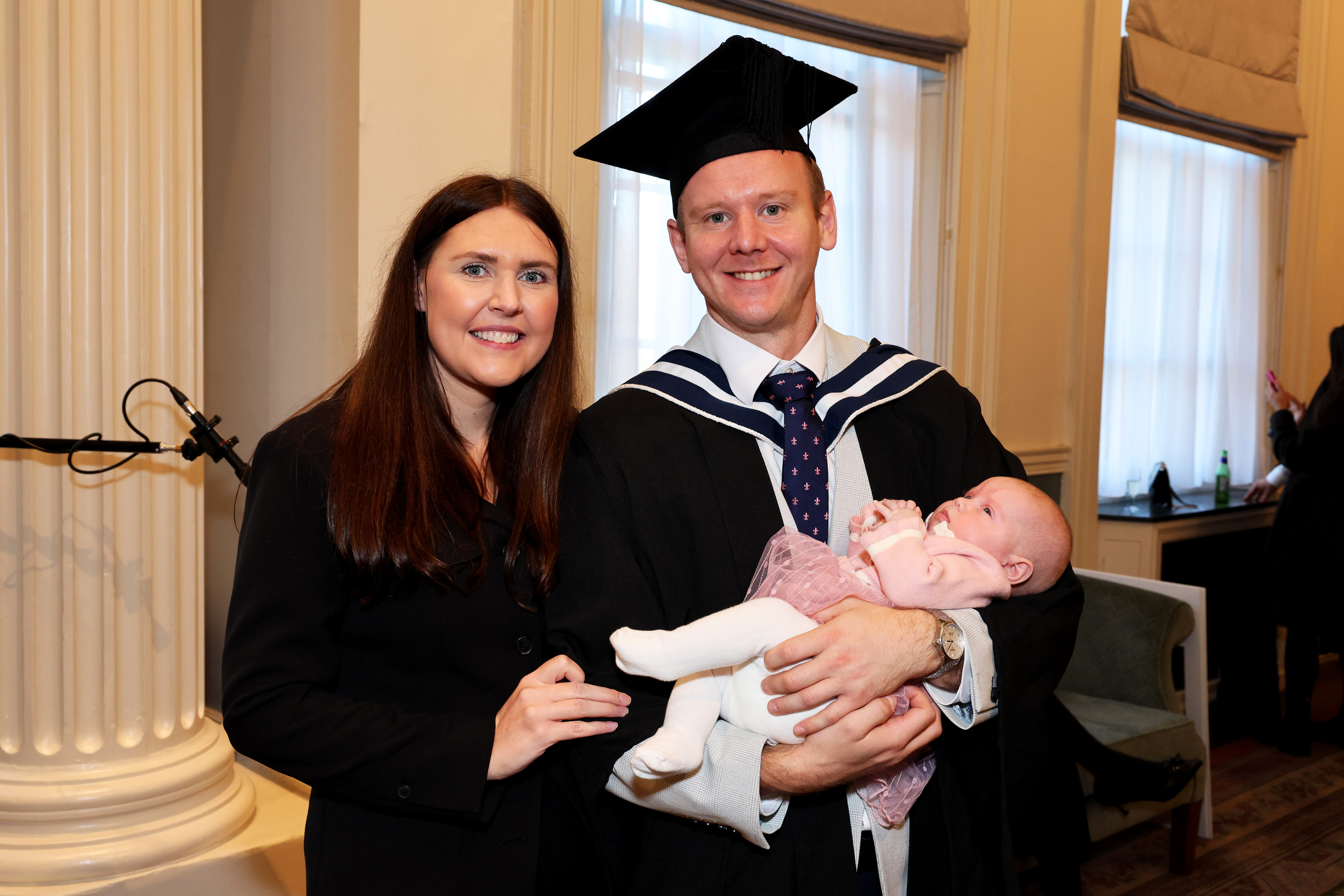 Man in graduation cap and gown poses for a picture with his female partner, holding a baby in his arms