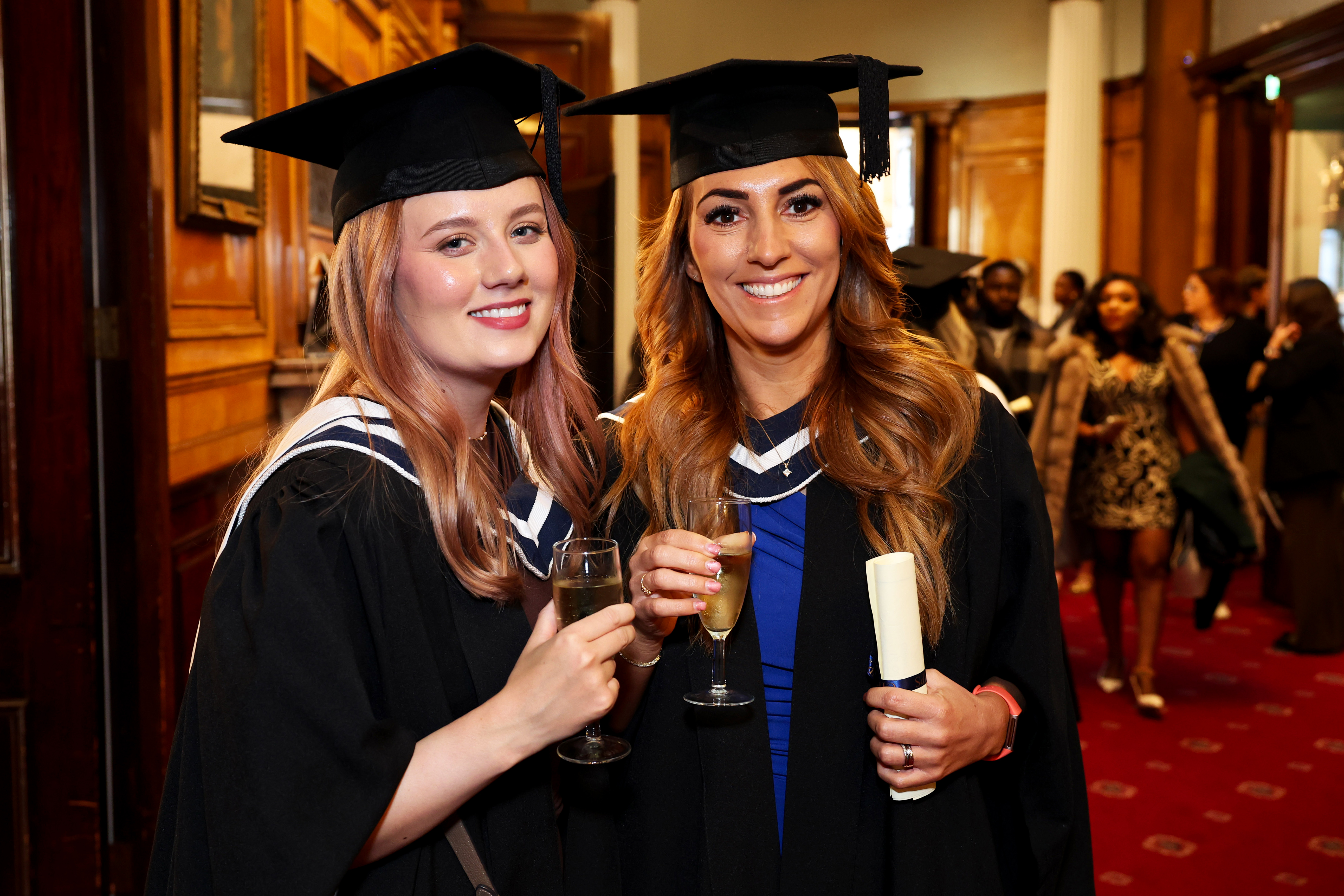 Two female graduates in cap and gown celebrating after the ceremony, holding drinks and showing off their diplomas