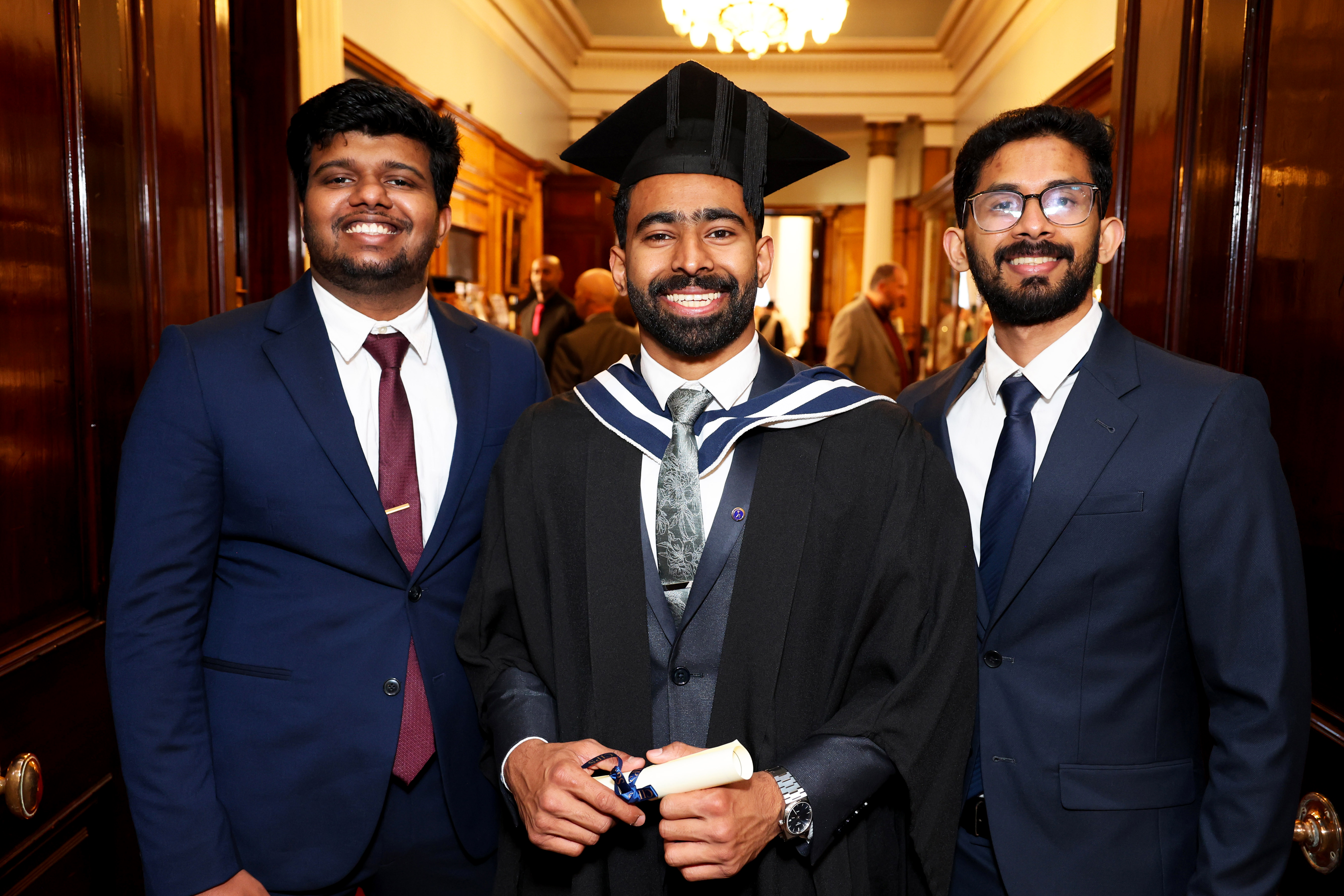 Male graduate in cap and gown posing for picture with two other men, friends or family