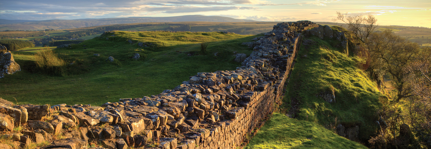 The stone ruins of Hadrian's Wall curve across the rolling green hills of the English countryside at sunset.