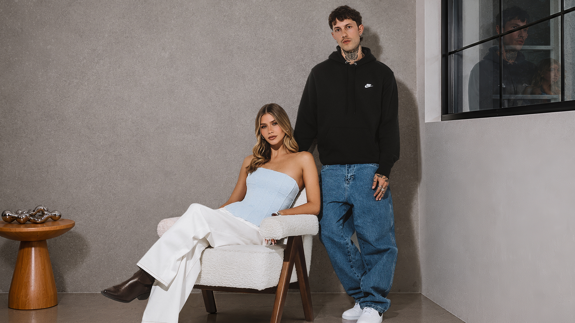 city beach, woman sitting on chair wearing white jeans strapless light blue top and black boots. male standing wearing black nike jumper blue jeans and white sneakers
