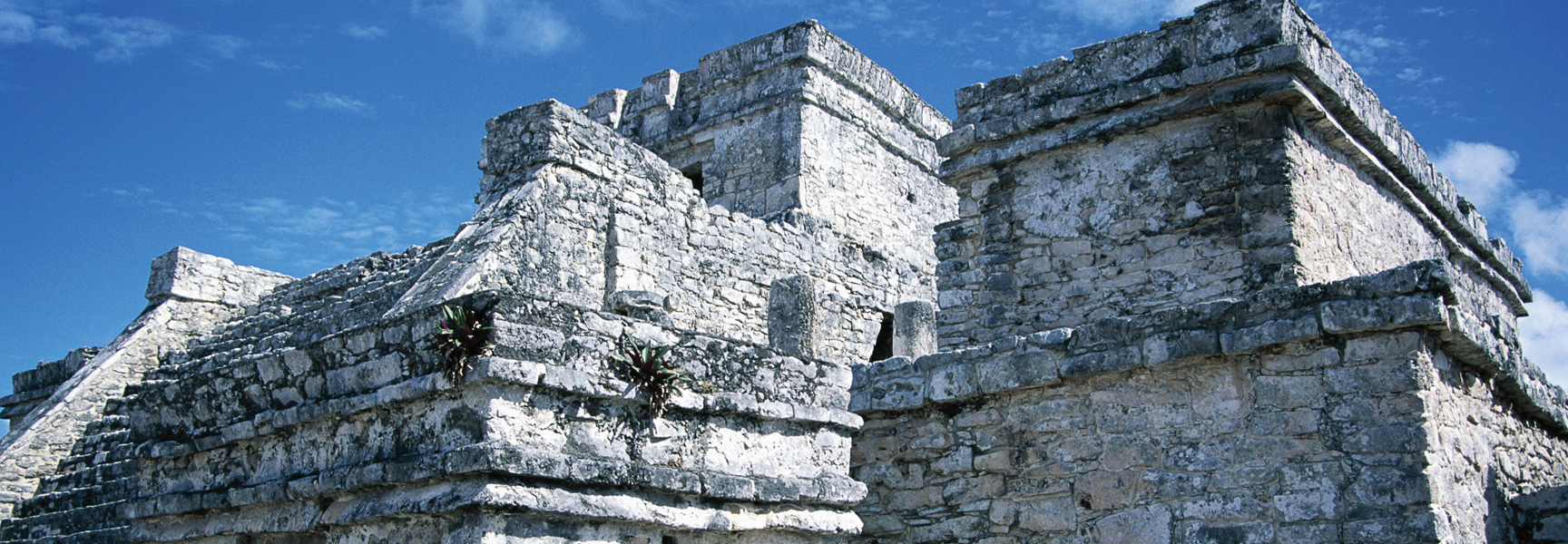 The top of ancient stone Mayan temple ruins in Belize/Guatemala are viewed from below against a bright blue sky.