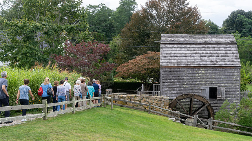 A tour group walks along a path beside the historic Dexter Grist Mill and its water wheel in Sandwich, Massachusetts.