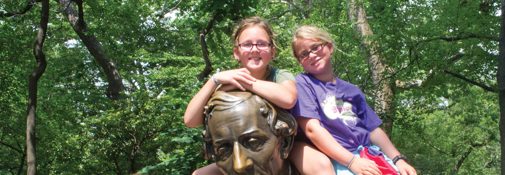 Two smiling children sit on a bronze bust of a man in a leafy New York park.