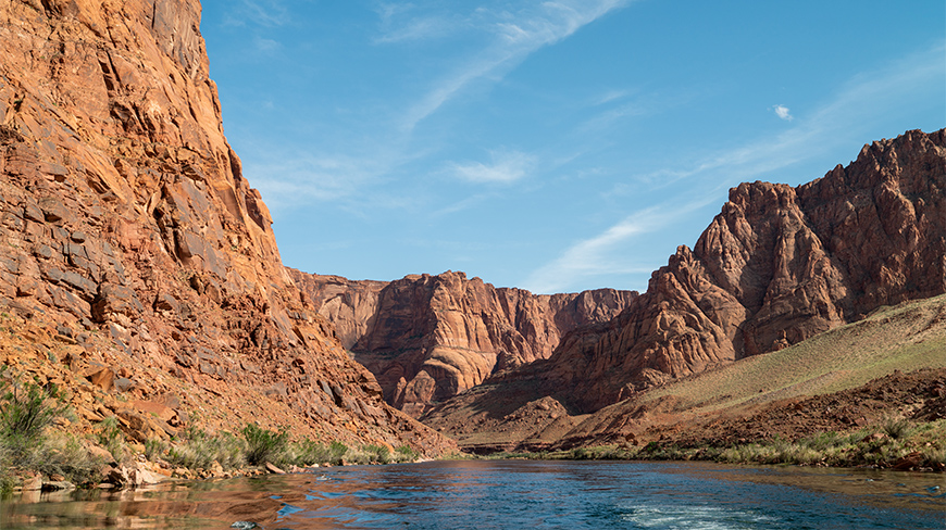 Rafting on the Colorado River through the steep, red-orange rock walls of the Grand Canyon in Arizona under a partly cloudy blue sky.