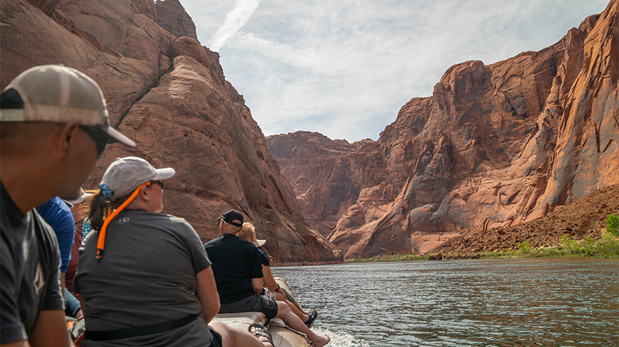 A group of people on a raft float down the Colorado River, surrounded by the massive red rock cliffs of the Grand Canyon in Arizona.