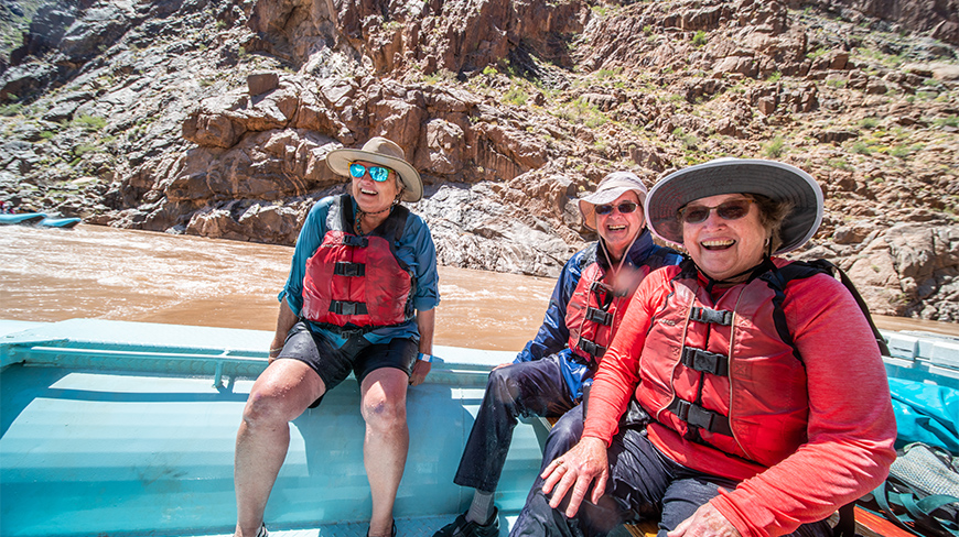 Three smiling women in life vests enjoy a sunny day rafting on the Colorado River in the Grand Canyon, Arizona.