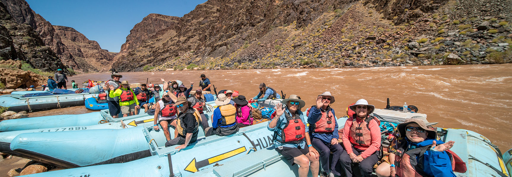 A group of people in life vests wave from their blue rafts on the Colorado River in the Grand Canyon, Arizona.
