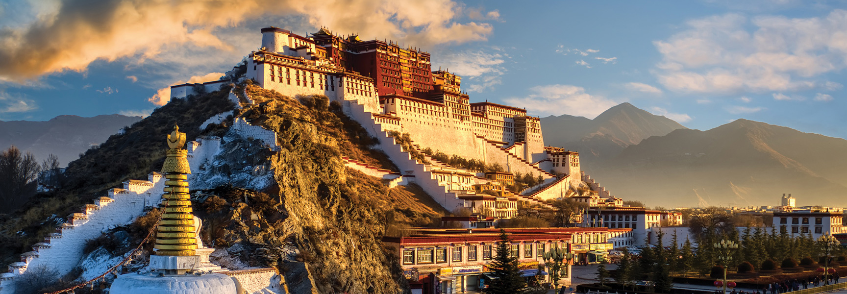The magnificent Potala Palace in Tibet sits atop a mountain, illuminated by the golden light of the setting sun with mountains in the background.
