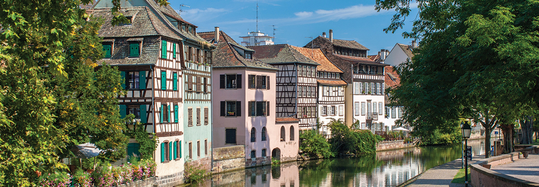 Colorful half-timbered buildings with vibrant shutters line a tranquil canal in France, their reflections shimmering on the water under a clear blue sky.