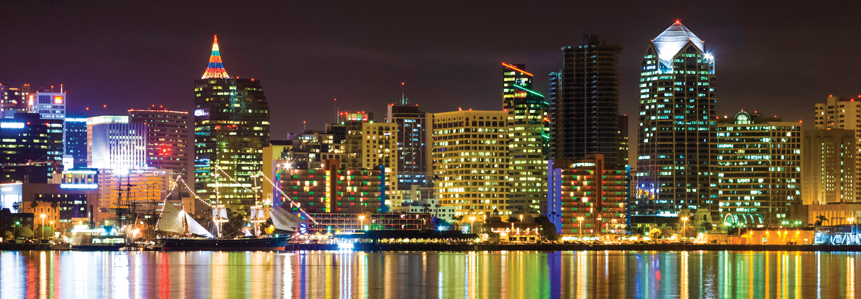 The San Diego, California skyline glows with colorful lights at night, reflecting on the water with a large sailboat docked in the foreground.