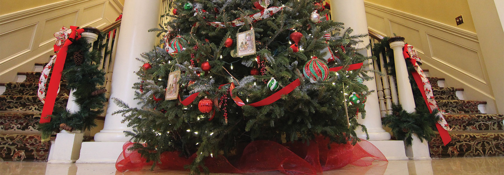 A large Christmas tree with red and green ornaments stands between two white pillars at the bottom of a grand staircase in Charleston, South Carolina.