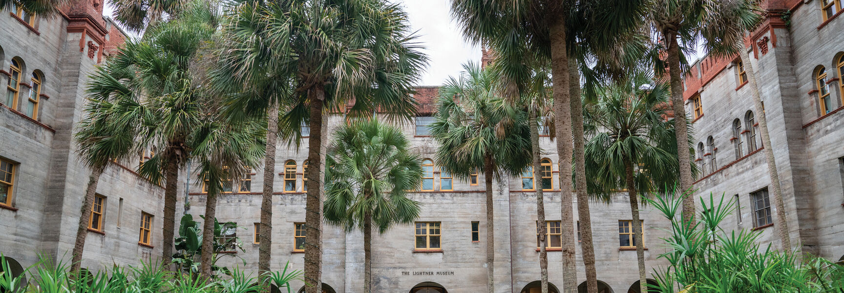 The lush courtyard of The Lightner Museum in St. Augustine, Florida, featuring palm trees, a fountain pond, and grand historic architecture.