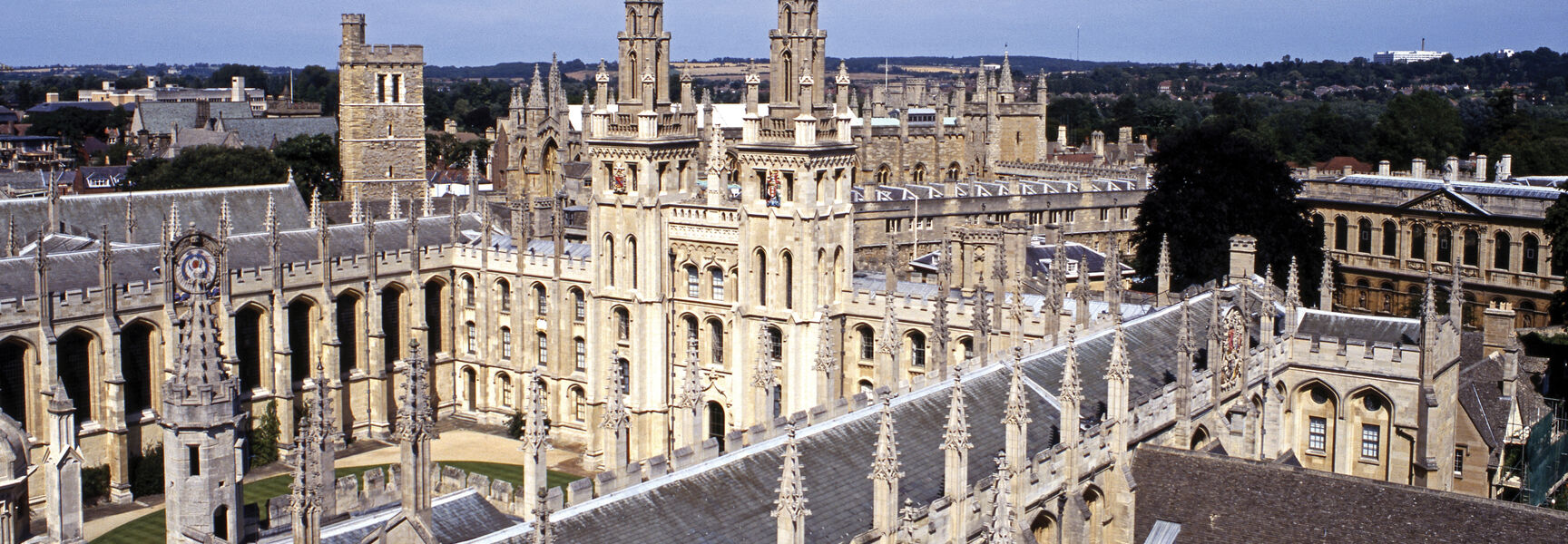 An aerial view of the historic stone buildings and Gothic spires of Oxford University in England under a clear blue sky.