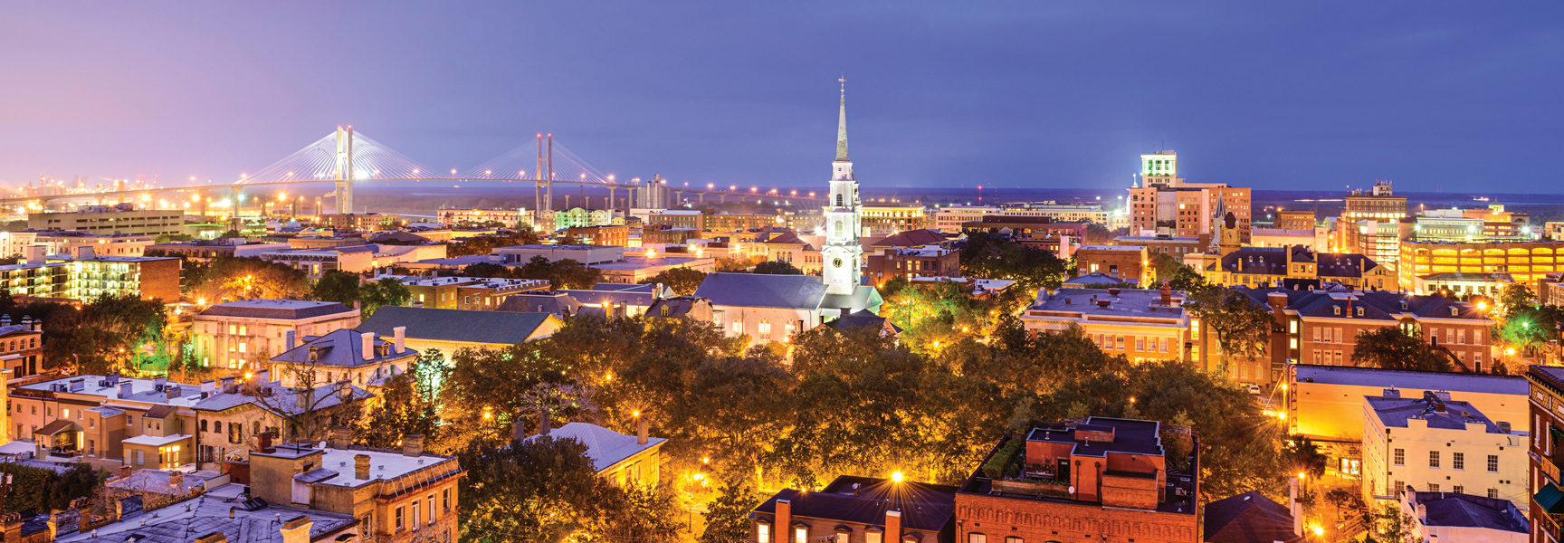 Aerial view of the Charleston, South Carolina skyline at dusk, featuring the illuminated city, a church steeple, and a bridge in the background.